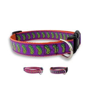 May include: Purple dog collar with a repeating pattern of green pickles on an orange background. The collar has a black plastic buckle and a silver metal D-ring.