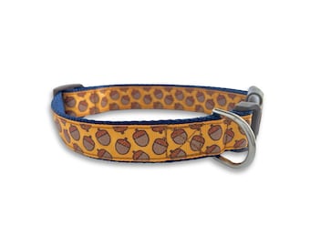 May include: A yellow dog collar with a navy blue trim and a pattern of brown acorns. The collar has a silver metal buckle and D-ring.