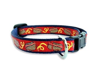 May include: A pet collar featuring a red ribbon overlay with a repeating pattern of brown hot dogs and yellow mustard swirls, stitched onto a navy blue nylon webbing base. The collar includes a black plastic side-release buckle and a silver metal D-ring.