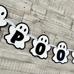 Op de afbeelding: Een witte spookbanner met zwarte letters die "SPOOKY" spellen. De banner is aan elkaar geregen met wit lint.