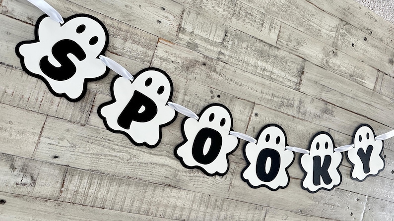 Op de afbeelding: Een witte spookbanner met zwarte letters die "SPOOKY" spellen. De banner is aan elkaar geregen met wit lint.