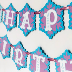 Pu&ograve; includere: Un festoso striscione con la scritta "HAPPY BIRTHAY" in lettere bianche. Ogni lettera &egrave; su un ritaglio a forma di fiocco di neve con un bordo azzurro e un centro viola. Lo striscione &egrave; infilato su un nastro rosa.