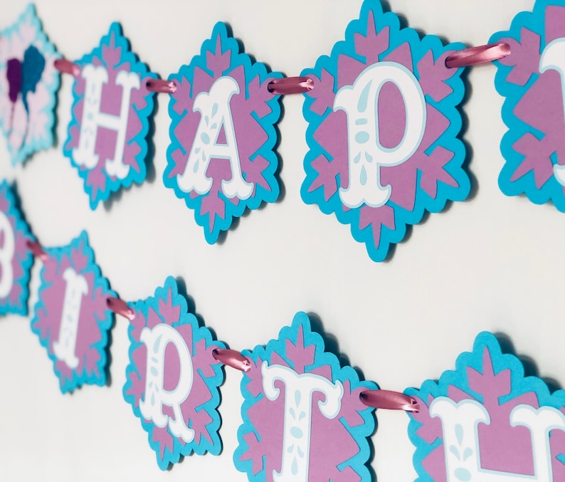 Pu&ograve; includere: Un festoso striscione con la scritta "HAPPY BIRTHAY" in lettere bianche. Ogni lettera &egrave; su un ritaglio a forma di fiocco di neve con un bordo azzurro e un centro viola. Lo striscione &egrave; infilato su un nastro rosa.