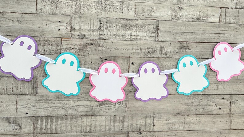 Op de afbeelding: Een Halloween-banner met zes witte spookuitsparingen. De spookjes zijn omrand met verschillende kleuren: paars, roze, turquoise en lichtblauw. De banner is aan elkaar geregen met wit lint.