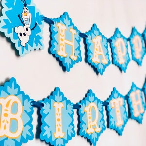 Pu&ograve; includere: Un banner di compleanno blu e bianco con le parole "HAPPY BIRTHDAY" e il personaggio Olaf. Lo striscione &egrave; composto da ritagli a forma di diamante con un design a fiocco di neve e lettere gialle.