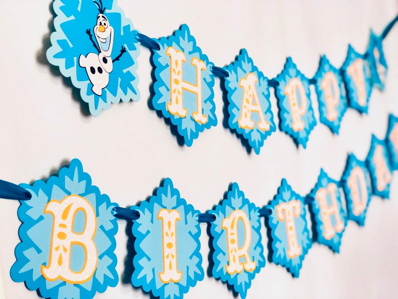 Pu&ograve; includere: Un banner di compleanno blu e bianco con le parole "HAPPY BIRTHDAY" e il personaggio Olaf. Lo striscione &egrave; composto da ritagli a forma di diamante con un design a fiocco di neve e lettere gialle.