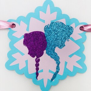 Pu&ograve; includere: Un banner a forma di fiocco di neve, con silhouette viola e blu glitterate su sfondo azzurro e rosa. Un nastro rosa &egrave; attaccato per appenderlo.