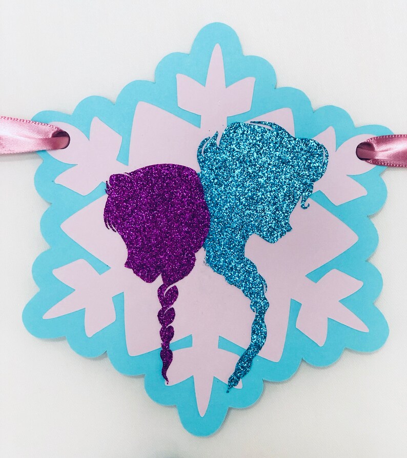 Pu&ograve; includere: Un banner a forma di fiocco di neve, con silhouette viola e blu glitterate su sfondo azzurro e rosa. Un nastro rosa &egrave; attaccato per appenderlo.