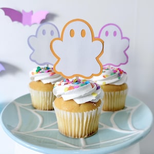May include: Four cupcakes with white frosting and colorful sprinkles sit on a light blue plate with a spiderweb design. Each cupcake has a paper ghost decoration. Purple and orange paper bats are in the background.