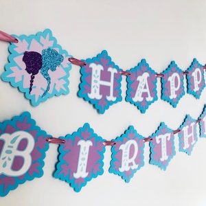 Pu&ograve; includere: Un festoso striscione di compleanno con un design a fiocco di neve. Lo striscione presenta le parole "HAPPY BIRTHDAY" in lettere bianche su ritagli a forma di diamante con uno schema di colori blu e viola. Un nastro rosa viene utilizzato per appendere lo striscione.