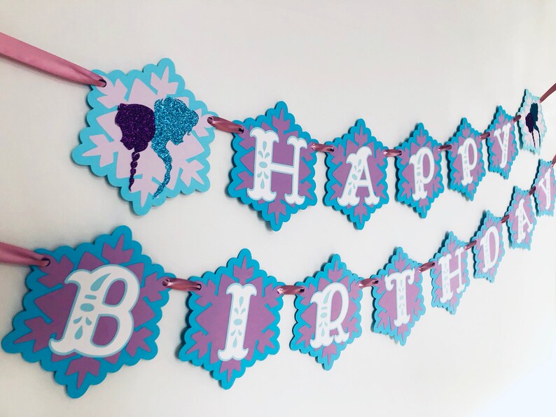 Pu&ograve; includere: Un festoso striscione di compleanno con un design a fiocco di neve. Lo striscione presenta le parole "HAPPY BIRTHDAY" in lettere bianche su ritagli a forma di diamante con uno schema di colori blu e viola. Un nastro rosa viene utilizzato per appendere lo striscione.