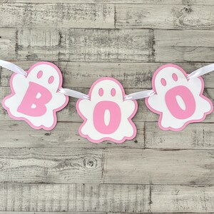 Op de afbeelding: Roze en witte spookbanner met het woord "BOO" erop. De spoken zijn wit met roze contouren en roze letters. De banner is geregen met wit lint.