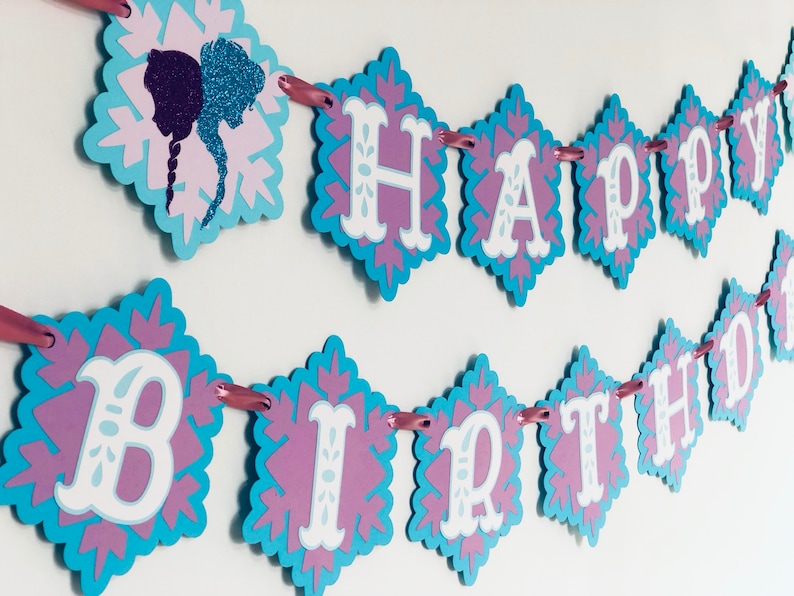 Pu&ograve; includere: Uno striscione decorativo con le parole "HAPPY BIRTHDAY" in bianco, su un disegno a fiocco di neve viola e blu. Lo striscione &egrave; infilato con un nastro rosa e presenta una silhouette di due figure.