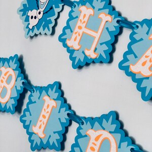 Pu&ograve; includere: Un banner di fiocchi di neve blu e bianchi con le parole "HAPPY BIRTHDAY" in lettere arancioni e bianche. Il banner include un'immagine di Olaf del film Frozen. Il banner &egrave; infilato con un nastro blu.