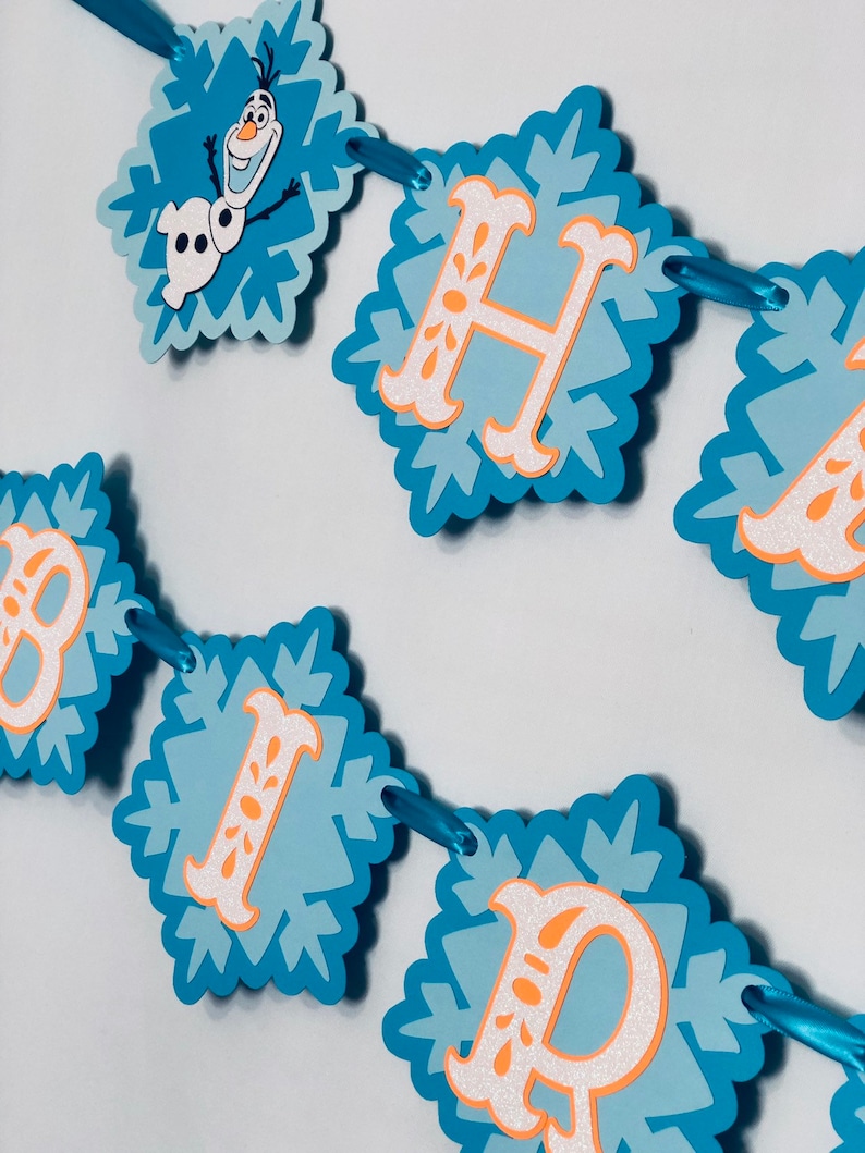 Pu&ograve; includere: Un banner di fiocchi di neve blu e bianchi con le parole "HAPPY BIRTHDAY" in lettere arancioni e bianche. Il banner include un'immagine di Olaf del film Frozen. Il banner &egrave; infilato con un nastro blu.