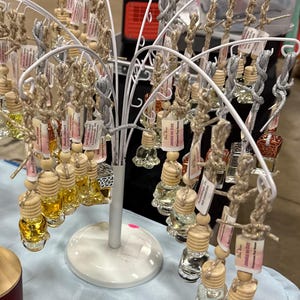 May include: A display of small glass bottles filled with various colored liquids, each topped with a wooden cap and hanging from a white metal tree-shaped stand. The bottles are suspended by twine and silver string, with small tags attached.