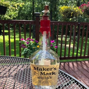 May include: A repurposed Maker's Mark whiskey bottle with a red wax seal and a copper-colored top. The bottle has a clear glass body with a label that reads "Maker's Mark Kentucky Straight Bourbon Whisky". The bottle is sitting on a metal table.