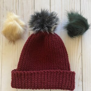 May include: A burgundy crocheted beanie with a black and white faux fur pom pom. Three additional pom poms are shown in the image: one beige, one black, and one green.