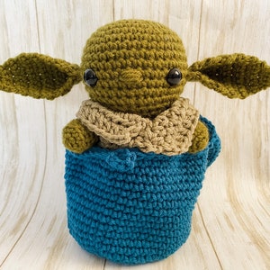 May include: A green crocheted baby Yoda doll wearing a brown crocheted scarf and sitting in a blue crocheted pot.