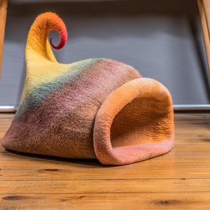 May include: A colorful felt cat bed shaped like a mushroom with a curled top. The bed is on a wooden floor with a stuffed squirrel and mushroom toy nearby.
