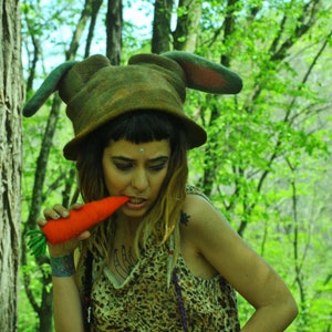 May include: A woman wearing a brown felt hat with bunny ears and a leopard print dress holds a carrot in her mouth. She is standing in a forest setting.