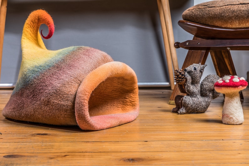 May include: A colorful felt cat bed shaped like a mushroom with a curled top. The bed is on a wooden floor with a stuffed squirrel and mushroom toy nearby.