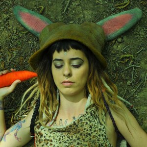 May include: A woman with long brown and blonde dreadlocks wears a green felt hat with pink bunny ears and a brown and white animal print top. She holds a bright orange carrot in her hand.
