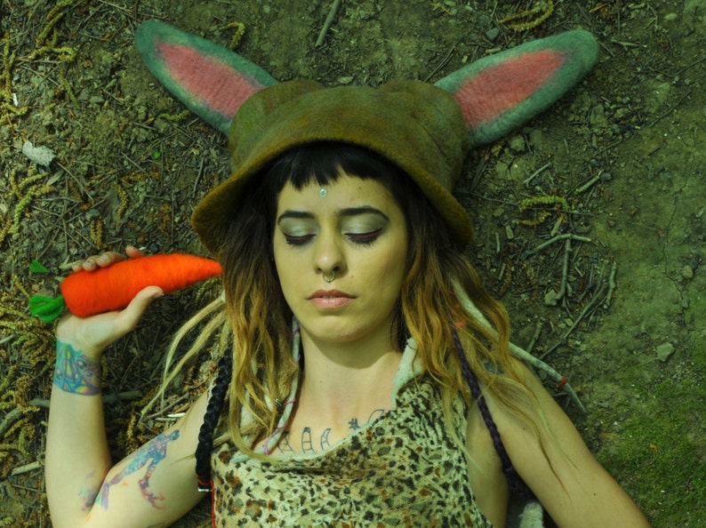 May include: A woman with long brown and blonde dreadlocks wears a green felt hat with pink bunny ears and a brown and white animal print top. She holds a bright orange carrot in her hand.