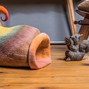 May include: A colorful felt cat bed shaped like a mushroom with a curled top. The bed is on a wooden floor with a gray felt squirrel and a red and white felt mushroom.