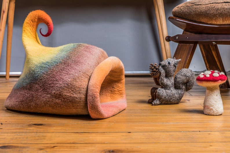 May include: A colorful felt cat bed shaped like a mushroom with a curled top. The bed is on a wooden floor with a gray felt squirrel and a red and white felt mushroom.