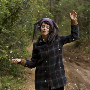 Puede incluir: Una mujer con una chaqueta de cuadros con capucha y un sombrero puntiagudo de colores. Est&aacute; de pie en un bosque con nieve cayendo a su alrededor.