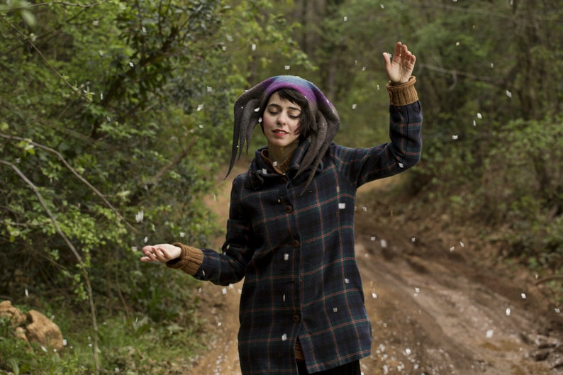 Puede incluir: Una mujer con una chaqueta de cuadros con capucha y un sombrero puntiagudo de colores. Est&aacute; de pie en un bosque con nieve cayendo a su alrededor.