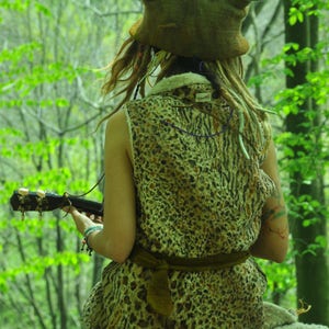May include: A person wearing a brown, furry hat with large ears sits on a rock in a forest. They are wearing a leopard print dress and holding a ukulele.