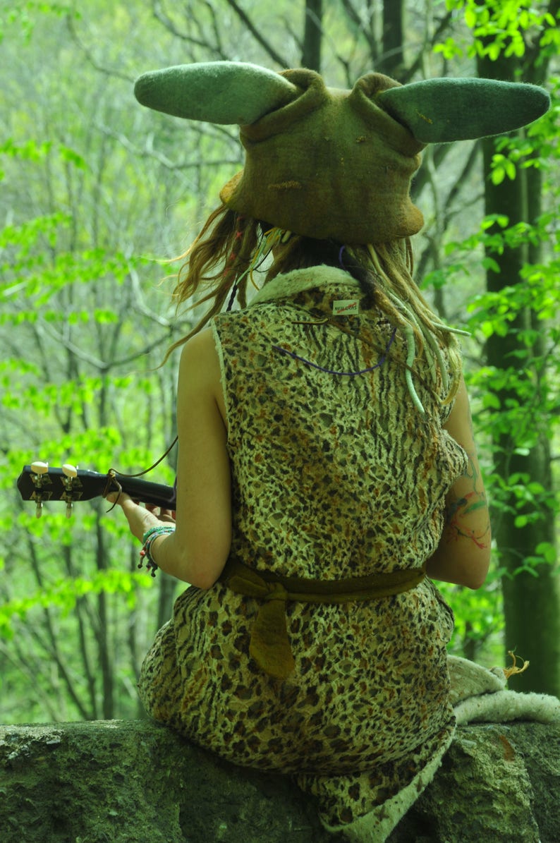 May include: A person wearing a brown, furry hat with large ears sits on a rock in a forest. They are wearing a leopard print dress and holding a ukulele.