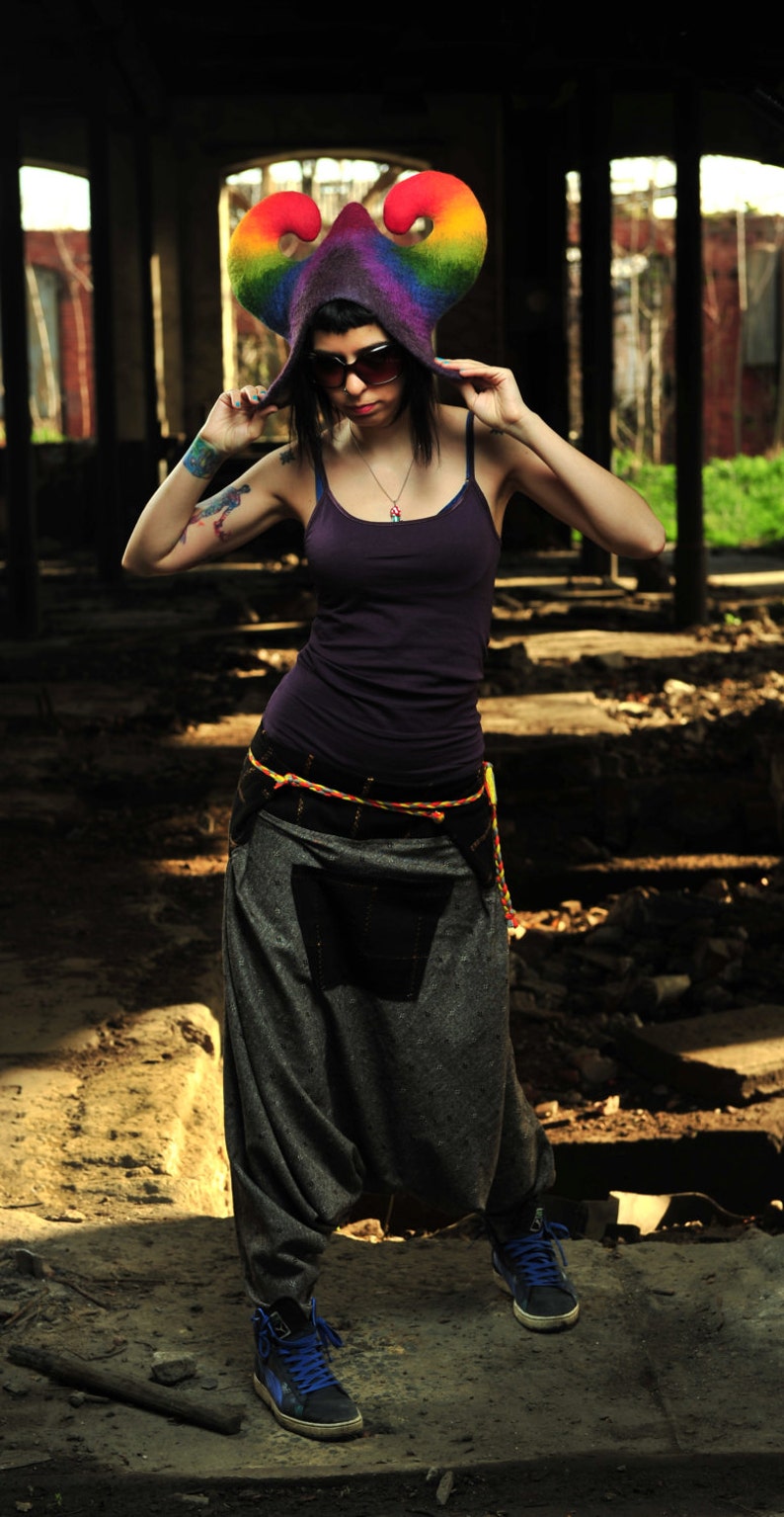 May include: A person wearing a rainbow-colored hat with a purple top and gray pants. The hat has a unique design with curved horns and a rainbow gradient. The outfit includes a dark purple tank top, gray harem pants, and blue sneakers.
