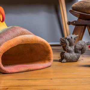May include: A colorful felt cat bed shaped like a mushroom with a curled top. The bed is on a wooden floor with a gray squirrel figurine and a red and white felt mushroom figurine.