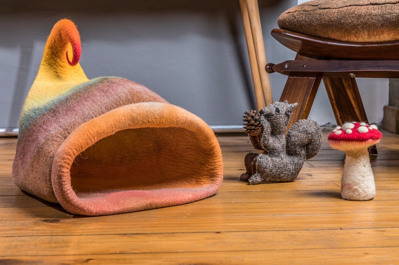 May include: A colorful felt cat bed shaped like a mushroom with a curled top. The bed is on a wooden floor with a gray squirrel figurine and a red and white felt mushroom figurine.