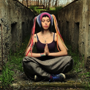 May include: A woman wearing a colorful hat with long braids sits cross-legged in a meditative pose. She is wearing a dark purple tank top and grey pants. The background is a concrete wall with a rough texture.