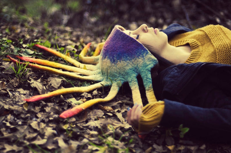 Op de afbeelding: Een vrouw draagt een kleurrijke, veelkleurige, octopusvormige hoed met lange, vloeiende tentakels. De hoed is gemaakt van vilt en heeft een regenboogkleurenschema. De vrouw ligt op haar zij met haar ogen dicht. De hoed is het middelpunt van de afbeelding.