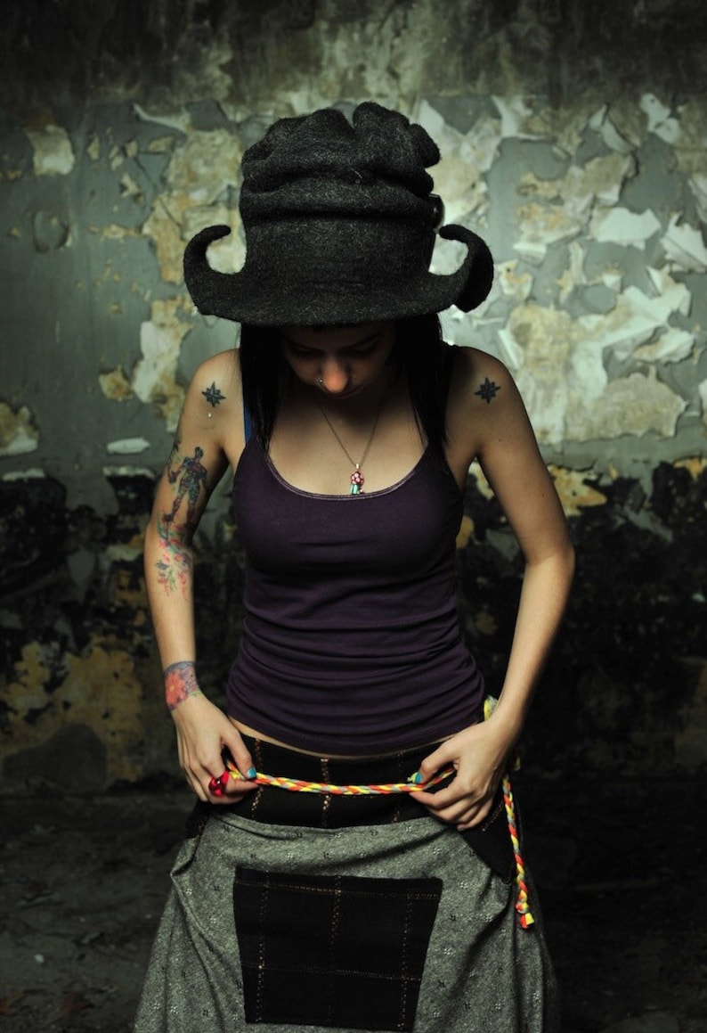 May include: A woman wearing a black hat with a unique design, a purple tank top, and a gray skirt with a black pocket. She is wearing a necklace with a turquoise pendant.