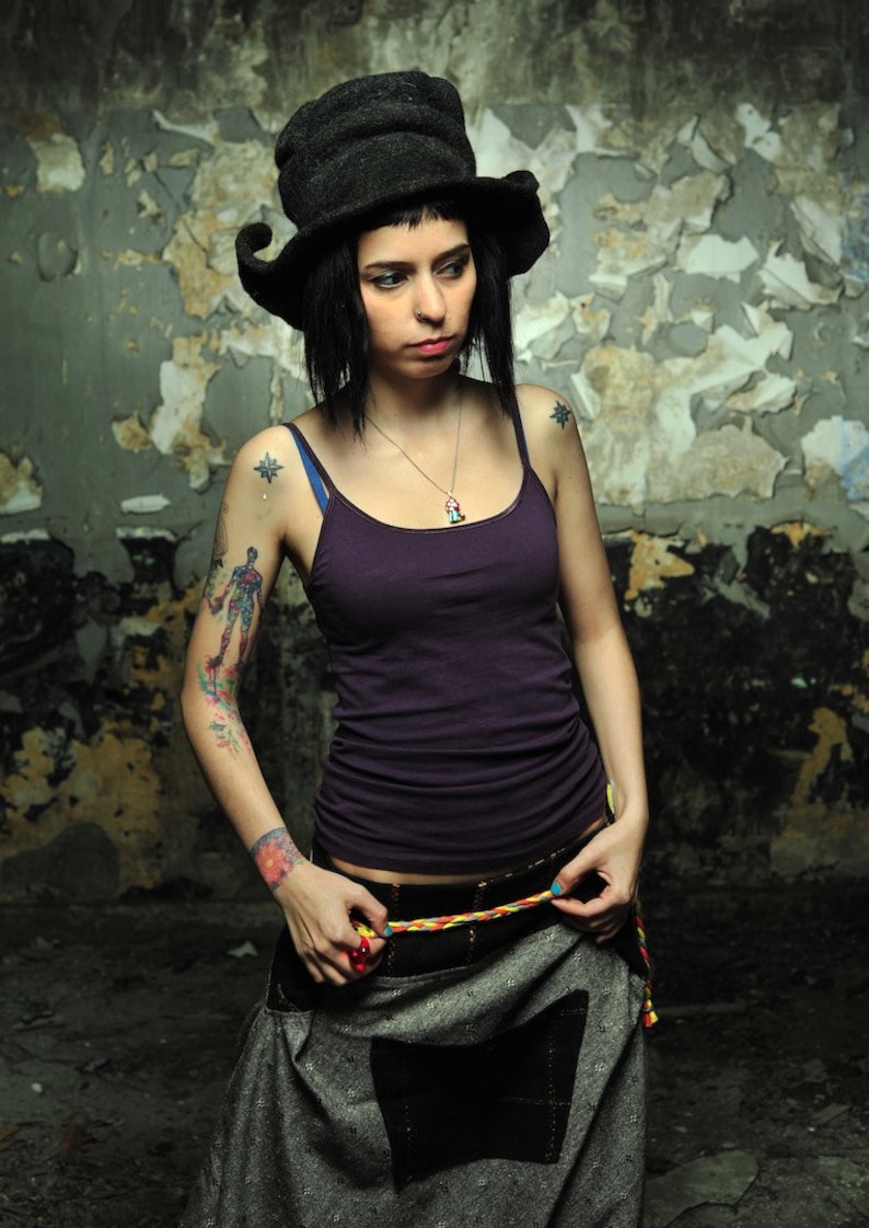 May include: A woman wearing a black hat with a wide brim, a purple tank top, and a grey skirt with a black pocket. She has tattoos on her arms and is wearing a silver necklace with a charm.