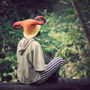 May include: A person wearing a colorful, horned hat sits in a forest setting. The hat is made of felt and has a red, orange, and yellow color scheme. The person is wearing striped pants and a gray sweater.
