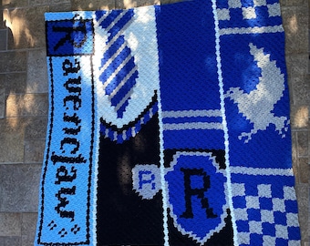 Puede incluir: Una manta de crochet azul, blanca y negra con el escudo de la casa Ravenclaw de Hogwarts. La manta presenta los colores de la casa Ravenclaw, un cuervo y la letra "R".