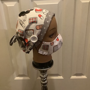 May include: A white and gray surgical scrub hat with a medical print featuring pills, bandages, and a syringe. The hat has a button closure and is displayed on a silver stand.