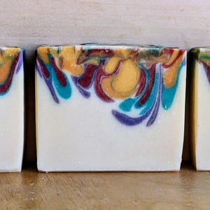 May include: Three rectangular bars of handmade soap. The soap has a white base with a colorful top layer featuring swirls of yellow, teal, burgundy, and purple. The soap is unscented and suitable for sensitive skin.