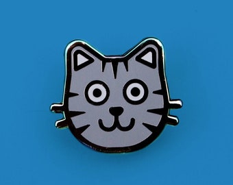 May include: A gray enamel pin with a cartoon cat face. The cat has black eyes, a black nose, and a black mouth. The cat's whiskers are also black.