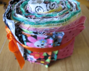 May include: A roll of colorful fabric scraps tied with an orange ribbon. The fabric scraps are arranged in a spiral pattern and feature various prints, including floral, skull, and polka dot designs.