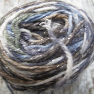 May include: A skein of yarn in a multi-colored pattern of white, brown, blue, and gray. The yarn has a subtle shimmer.