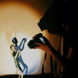 May include: A person taking a photograph of a ceramic figurine of a person wearing a green and yellow patterned shirt. The figurine is standing on a small platform.