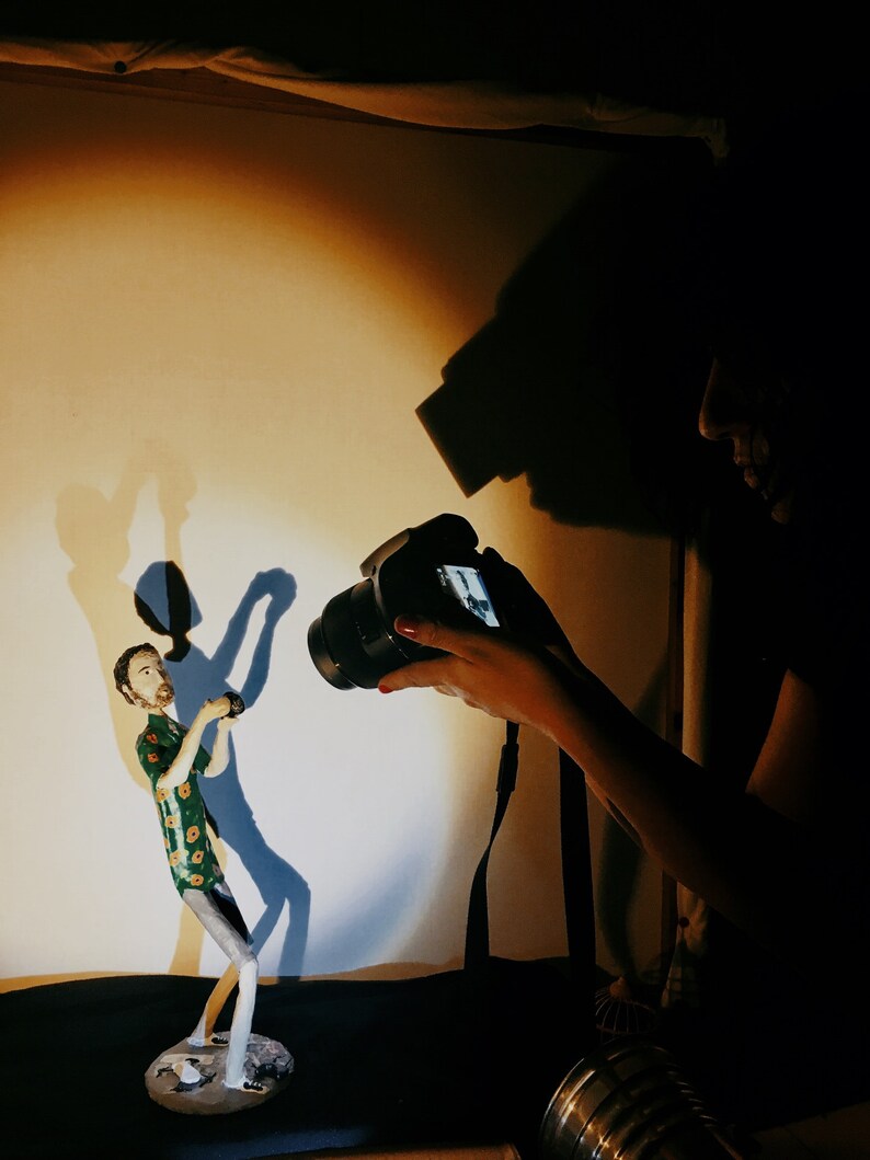 May include: A person taking a photograph of a ceramic figurine of a person wearing a green and yellow patterned shirt. The figurine is standing on a small platform.